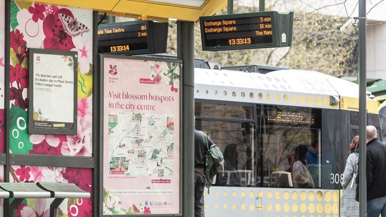The Bloomtown Map is displayed at a tram stop at St Peter's square in Manchester. Behind is a tram which is decorated with pink blossom and leaves.